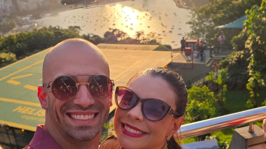 Casal no Morro da Urca ao pôr do sol, com helicóptero sobrevoando a enseada de Botafogo